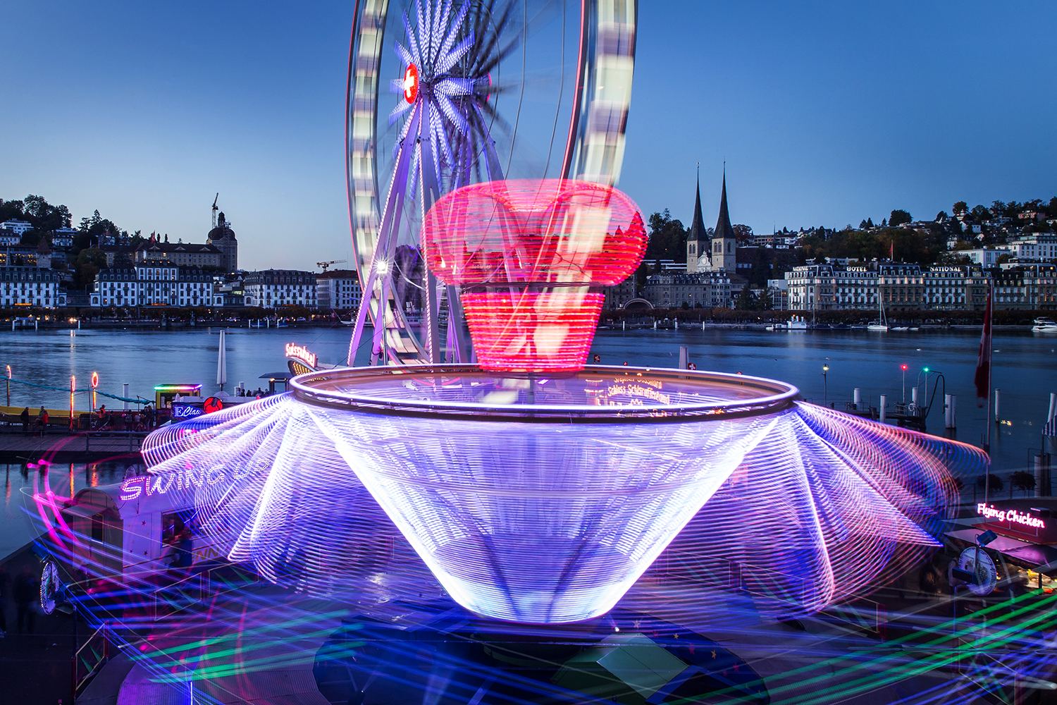 Lichtspuren eines Fahrgeschäfts vor Riesenrad und Hofkirche an der Luzerner Määs in der blauen Stunde